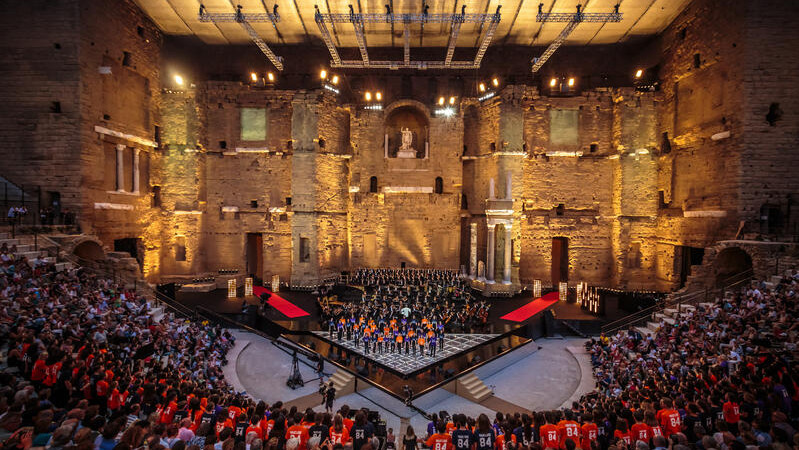 Photo prise depuis les gradins à la nuit tombée. Sur la scène au premier plan, l'orchestre et à l'arrière, on aperçoit le chœur